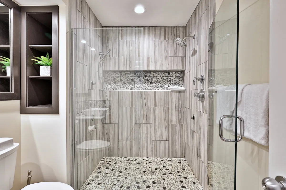 A modern bathroom featuring a glass shower enclosure with textured wall tiles, a stone floor, and a built-in shelf with decorative plants. The design is clean and serene, evoking a spa-like experience.
