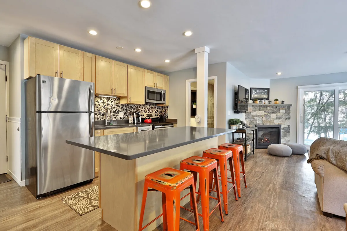 This angle highlights the kitchen island with stools, stainless steel appliances, and a stone fireplace, set off by a decorative backsplash. The communal feel is enhanced by the arrangement of seating and the prominence of the fireplace, emphasizing spaces for cooking, casual dining, and gathering.