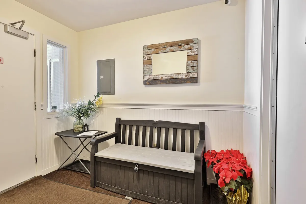 A cozy, shared lobby or waiting area featuring benches, potted flowers, a side table, and a mirror. The space feels inviting and communal, suggesting a place to relax, greet guests, or make social connections before and after exploring the area.