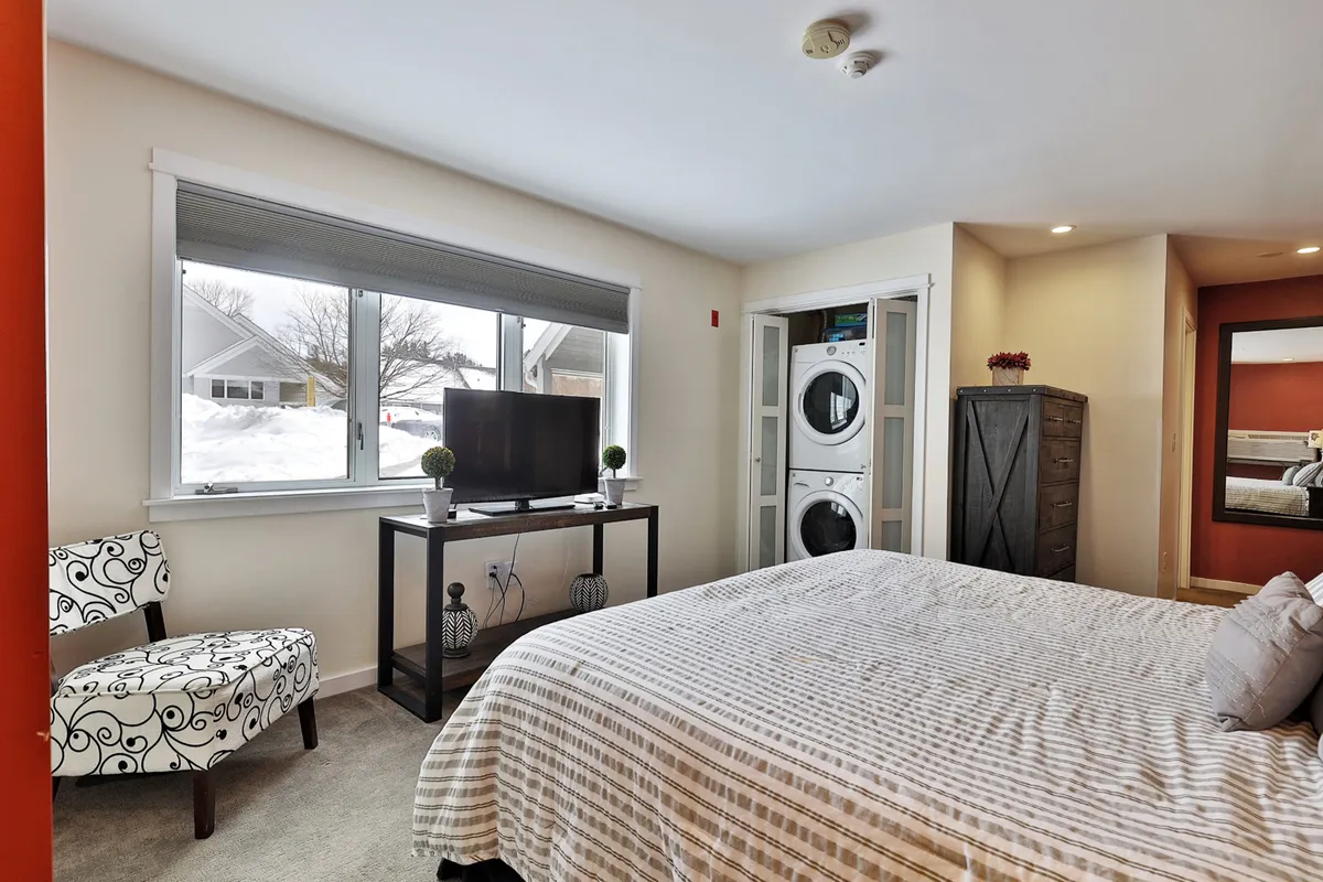 A plush bed is framed by a window, TV stand, decorative chair, and mirror, suggesting a warm spot for relaxation. The space looks inviting for couples to unwind with a movie or enjoy a quiet coffee by the window, with soft lighting enhancing the comforting mood.