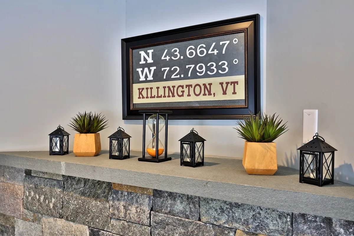 A cozy interior vignette features a decorative sign with the coordinates for Killington, VT, accompanied by soft lantern lighting and succulent plants on a mantel. The scene radiates a sense of place and intimacy, inviting guests to feel at home and connect with the mountain setting.