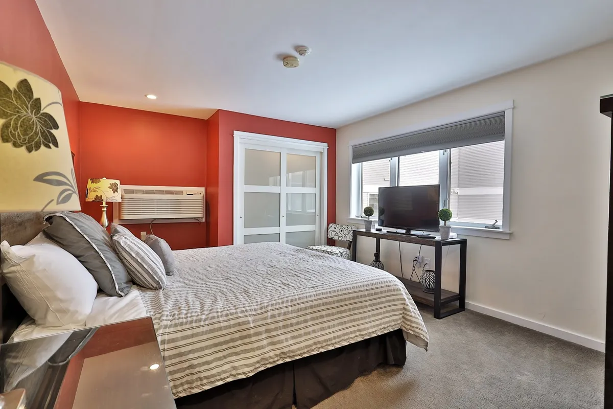 A cozy bedroom with a comfortable bed, a flat-screen smart TV mounted opposite the bed, and a window letting in natural light. The ambiance is calm and inviting, perfect for relaxation and privacy.