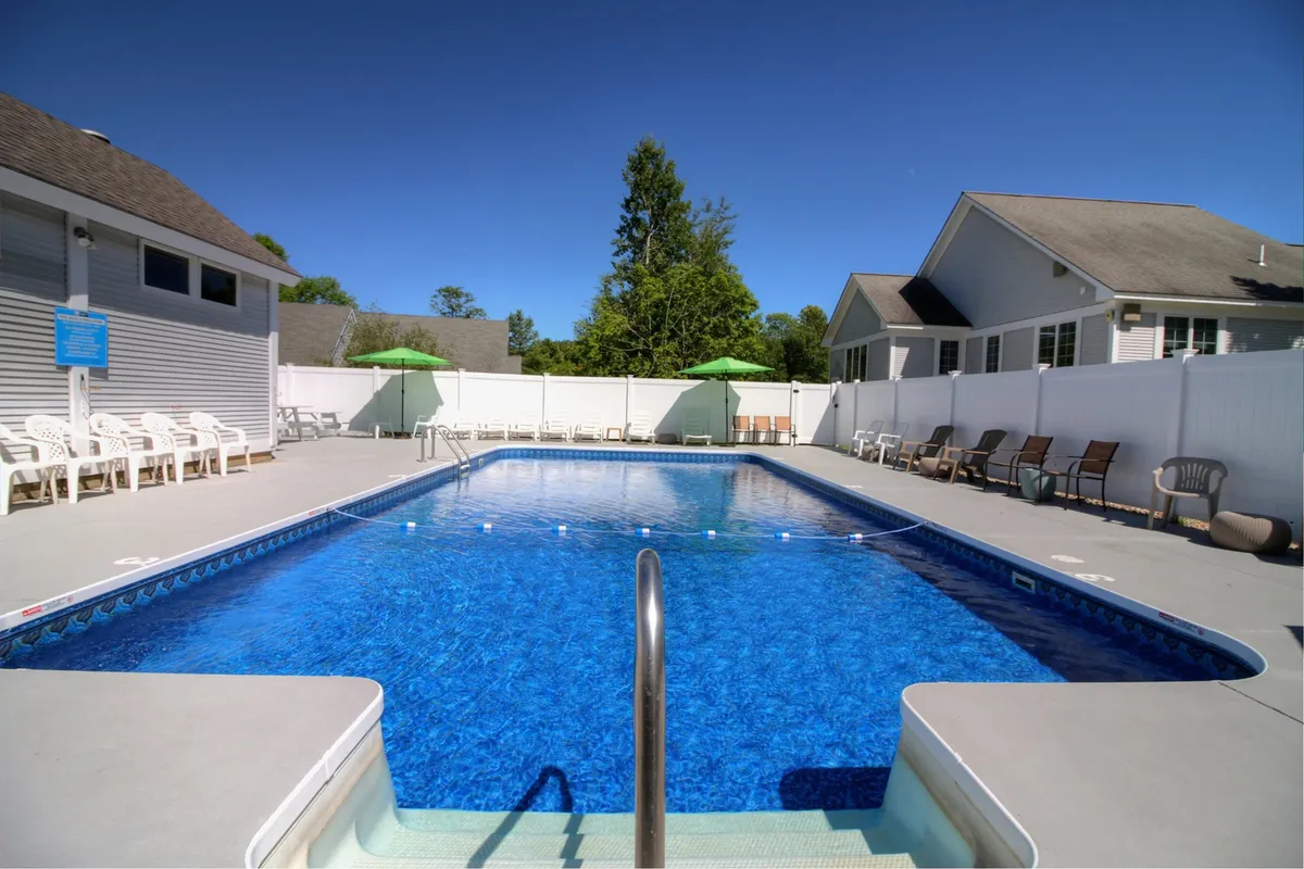 This image displays a vibrant outdoor pool area surrounded by lush trees, with sun loungers and green umbrellas set up for relaxation. The tranquil pool is inviting, and the scene radiates summer leisure, suggesting the perfect spot for a refreshing swim or sunbathing after a day of hiking or golf.