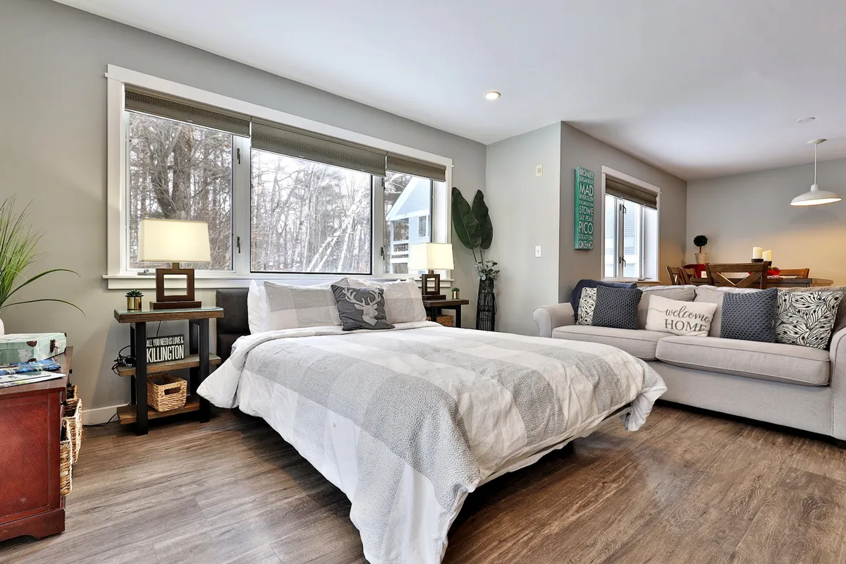 A spacious sleeping area featuring a bed with gray and white bedding and a sofa with decorative pillows, bathed in ample natural light from large windows. The background shows a dining area, suggesting a multi-functional suite that can comfortably accommodate groups or families.