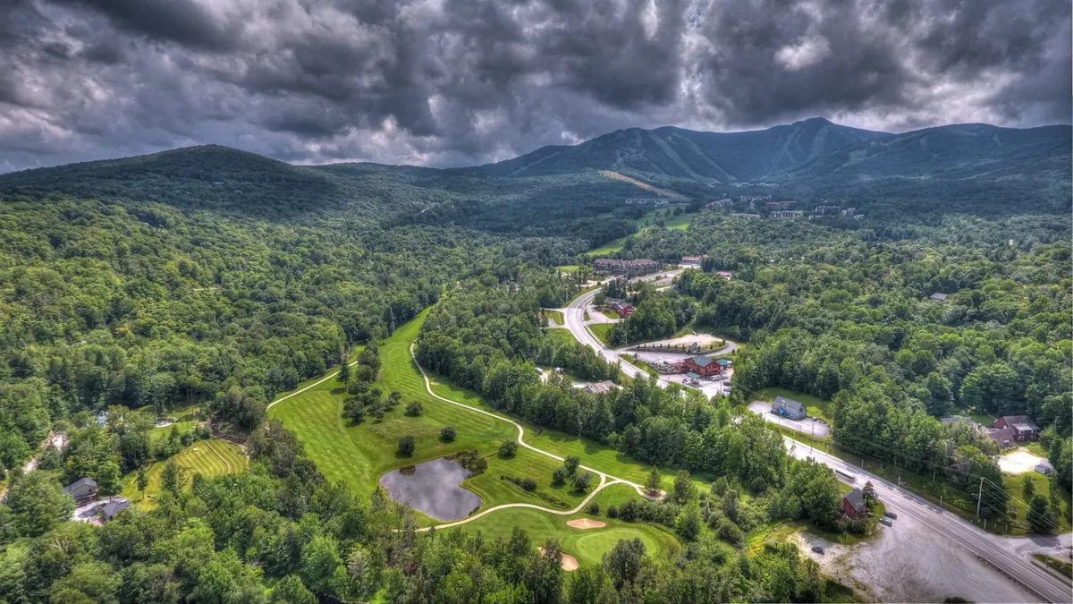 An expansive view encompasses a rolling golf course, a pond, dense forests, and towering mountains in the background. The peaceful landscape showcases both the golfing and hiking opportunities available in the area, with the natural beauty of Vermont in full display.