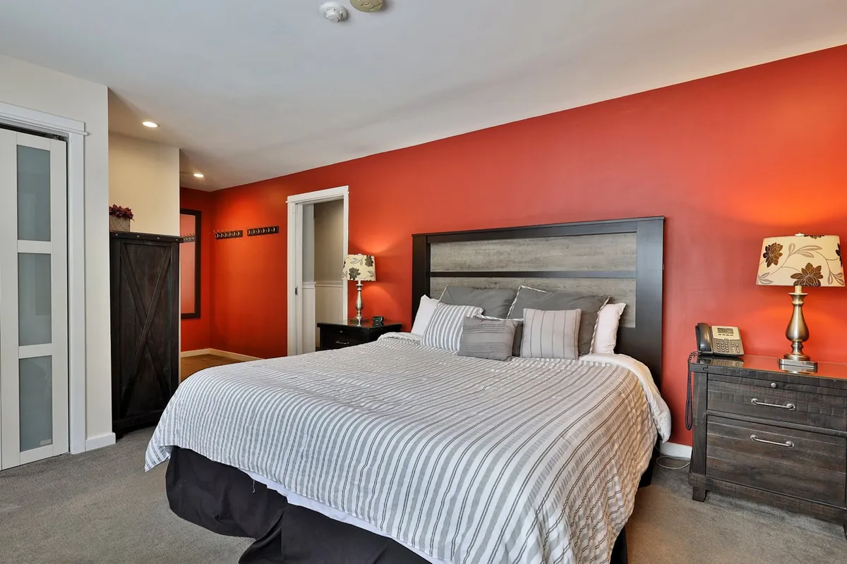 A restful bedroom with a comfortable bed, red accent wall, soft lighting from a bedside lamp, a nightstand, and a dresser. The ambiance is calm and restful, inviting relaxation and quiet moments.