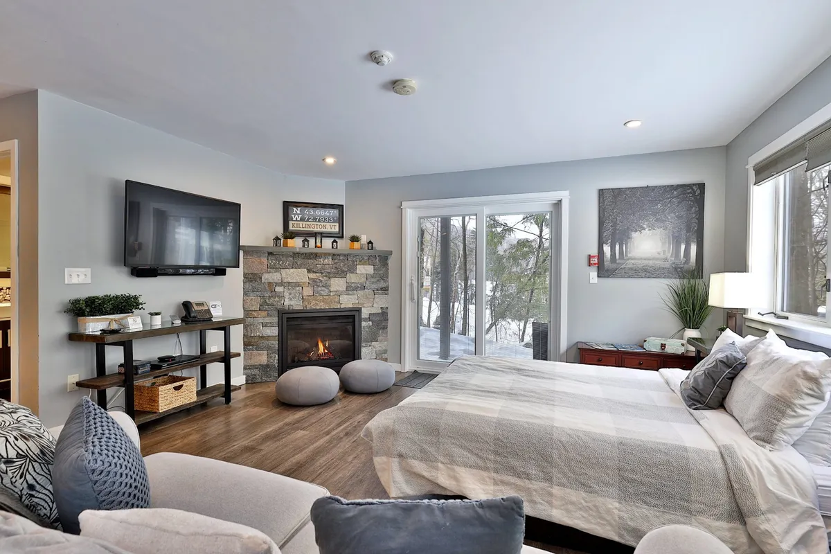 The image showcases a cozy living space with a crackling fireplace, plush bed, large TV, tasteful artwork, and a sliding glass door framing a snowy mountain view. The scene evokes comfort and relaxation, inviting guests to unwind after a day on the slopes.