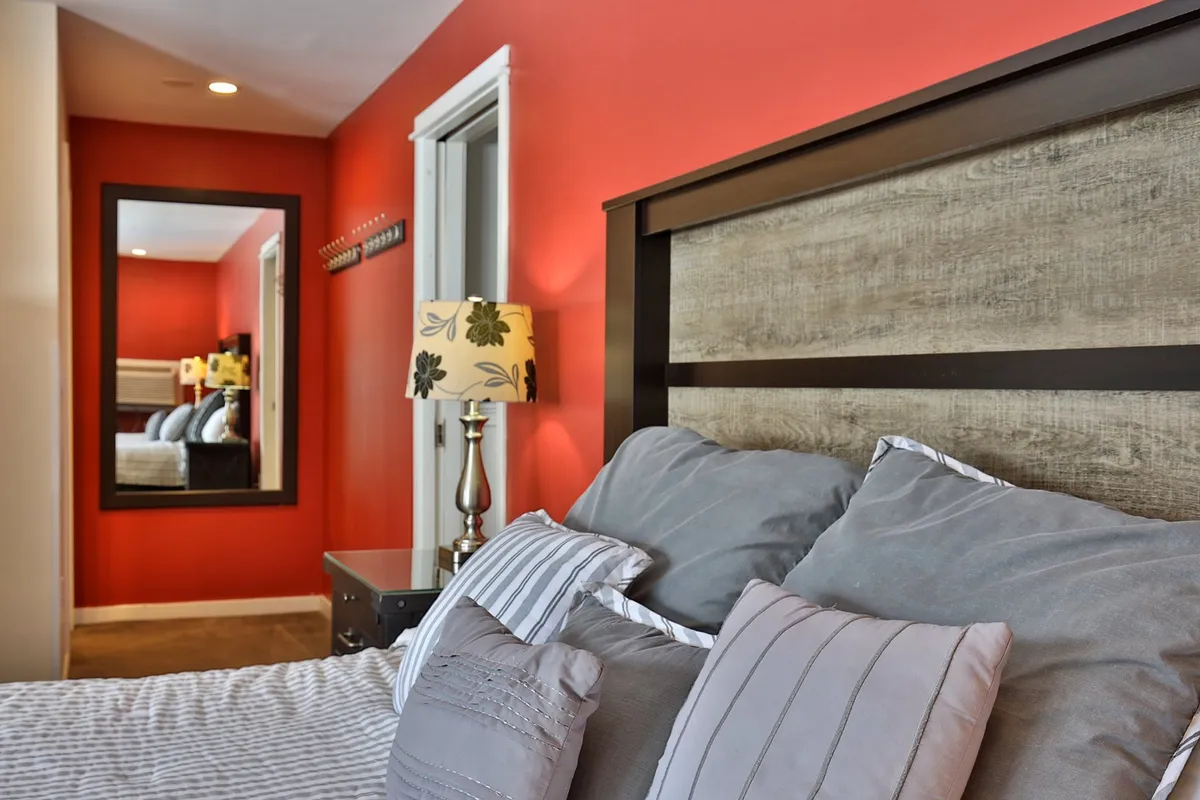 A tranquil bedroom scene with a well-dressed bed, decorative pillows, a warm lamp, and a mirror reflecting soft morning light. The ambiance is calm and private, ideal for romantic mornings or peaceful evenings.