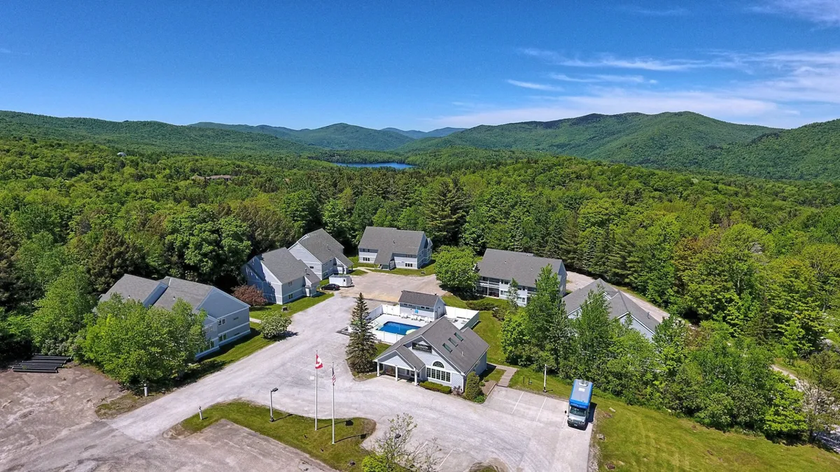 A wide exterior shot of the swimming pool with surrounding trees, a mountain backdrop, and the main building visible. The scene captures both the communal and scenic aspects of the property, highlighting its location amidst Vermont’s natural landscape and its inviting outdoor amenities.