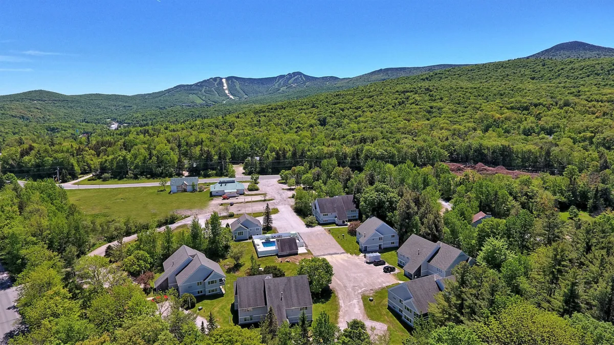 A wide-angle exterior shot features a cluster of houses nestled in the mountains, with a swimming pool in the foreground and trails winding into the landscape. The scene conveys a welcoming resort community surrounded by nature, ideal for group or family retreats.